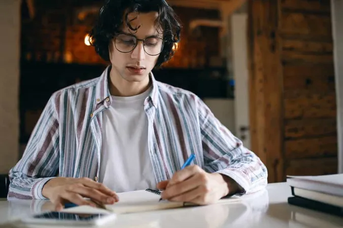 Focused Male Student Glasses Desk Making Notes Copybook Putting Down Thoughts Ideas Preparing Test Exam X - General Achievement Test study guide